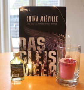Das Gleismeer - Miéville, Foto © Eva Bergschneider, Cover © Heyne Verlag, schwarzes Cover mit Engelsflügel mit Zahnrad, Bucht steht auf Esszimmertisch mit Kerze und Met-Fläschchen.