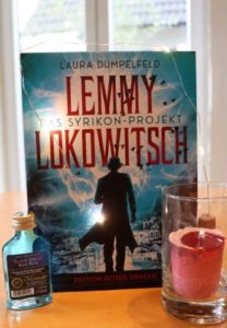 Lemmy Lokowitsch-Das Syrikon-Projekt - Laura Dümpelfeld © Eva Bergschneider, Cover © Edition Roter Drache, blaues Cover mit Mann mit Hut vor Stadt-Silhouette, Bucht steht auf Esszimmertisch mit Kerze und Met-Fläschchen.